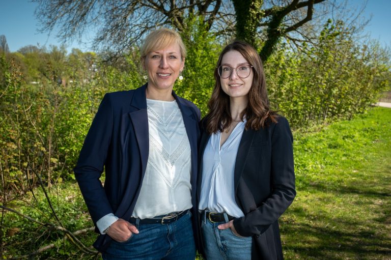 Zwei Frauen stehen lächelnd vor einem grünen Hintergrund, beide in Blazern gekleidet.