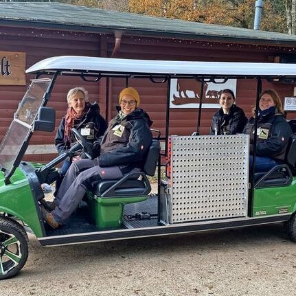 Vier Personen sitzen in einem grünen Fahrzeug vor einer Hüttenstruktur im Wald.