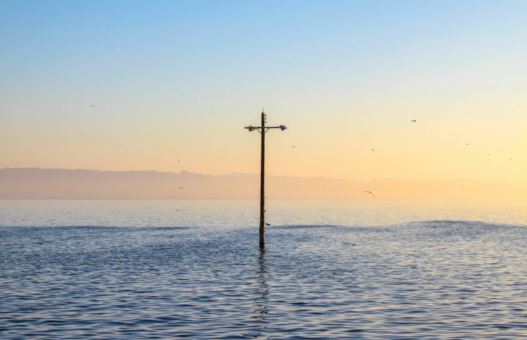 Überflutungsvorsorge Holzkreuz, das aus ruhigem Wasser ragt, umgeben von einem sanften Farbverlauf am Himmel.