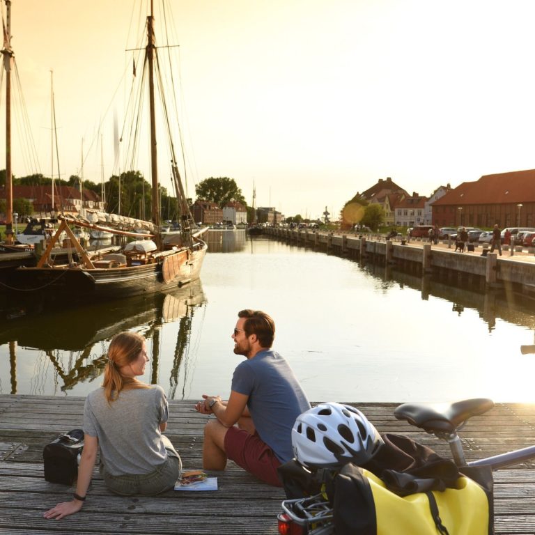 Paar sitzt am Hafen, Blick auf Boote und ruhiges Wasser bei Sonnenuntergang.