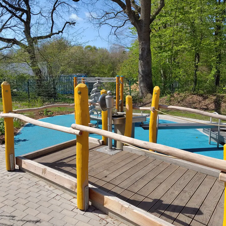 Spielplatz mit Holzplattform, blauer Fläche und grüner Umgebung.