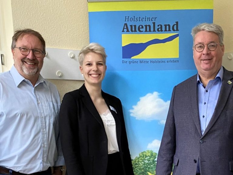 Drei Personen stehen vor einer Landschaft mit Wasser und Stadtansicht im Hintergrund.