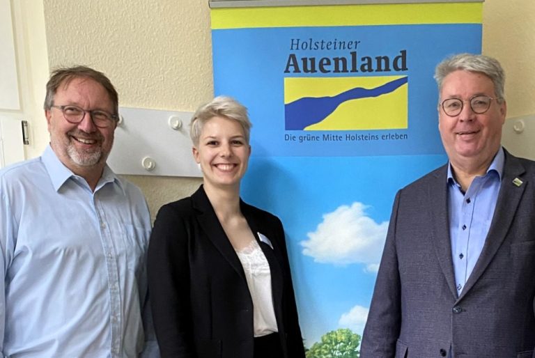 Drei Personen stehen lächelnd vor einer Landschaft mit Wasser und Stadt im Hintergrund.