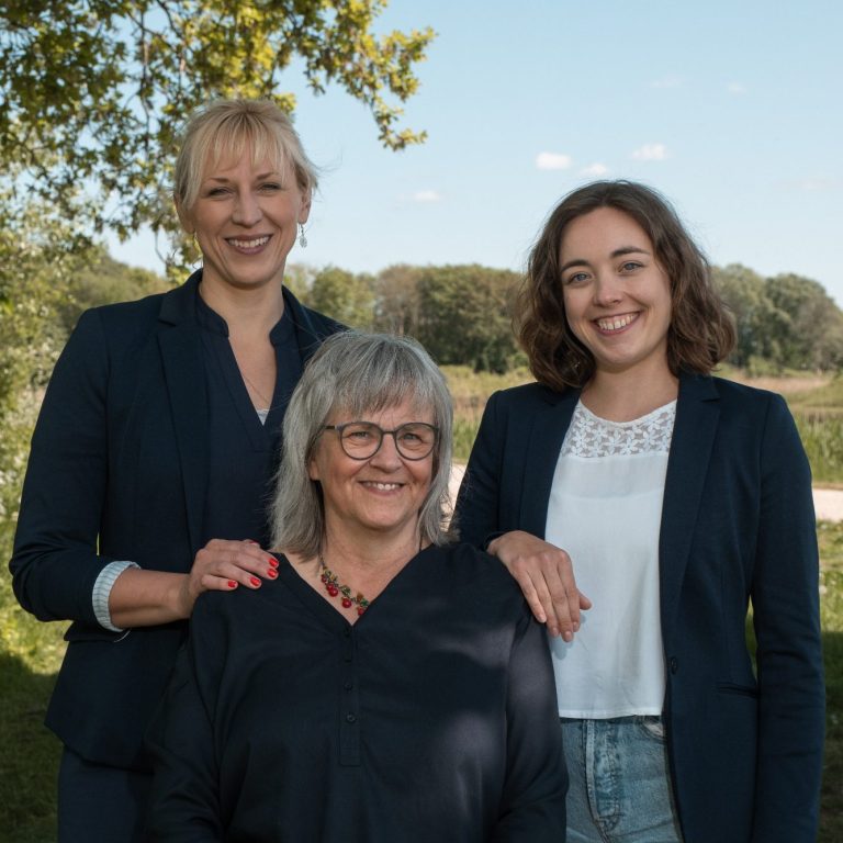 Drei Frauen stehen lächelnd im Freien, umgeben von grüner Natur.