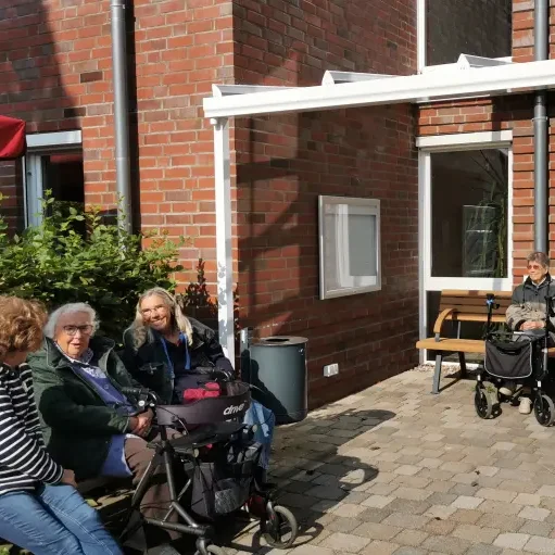 Ältere Menschen sitzen im Freien auf Bänken und Rollstühlen vor einem Gebäude.