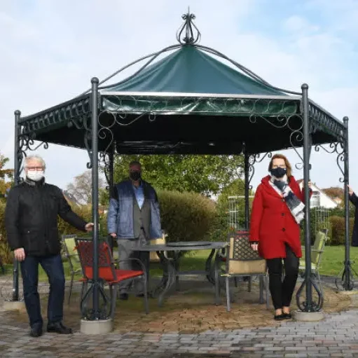 Personen in Maske stehen unter einem Pavillon im Freien, umgeben von Grün.