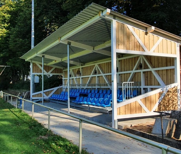 Überdachte Sitzgelegenheit auf einem Sportplatz mit blauen Sitzen und Holzstapel.