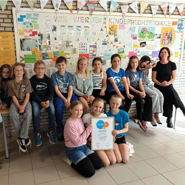 Gruppe von Kindern und Erwachsenen posiert vor einer bunten Wand mit Informationsmaterialien.