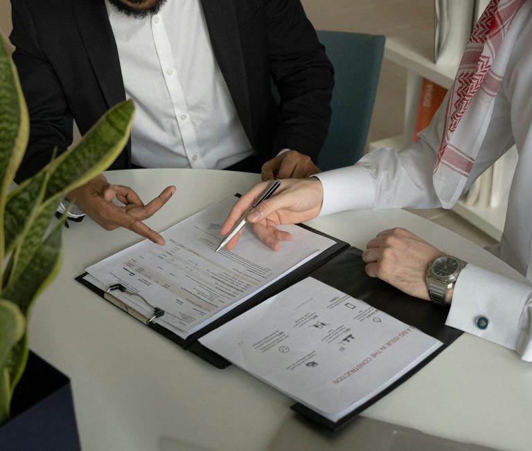 Zwei Personen in Anzügen besprechen Dokumente am Tisch, eine Hand zeigt auf die Papiere.