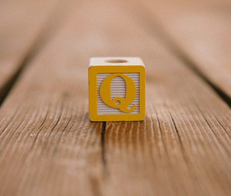 Holzblock mit einem gelben "Q" auf der Vorderseite, auf einer Holzoberfläche platziert.