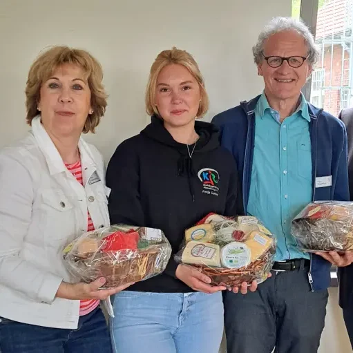 Drei Personen halten Geschenkkörbe, lächeln und stehen in einem hellen Raum.