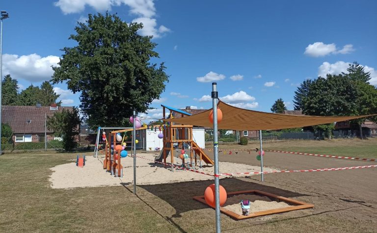 Spielplatz mit Klettergerüst, Rutsche, Sandkasten und Sonnenschutz unter blauem Himmel.