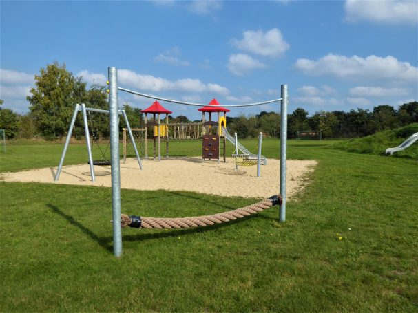 Spielplatz mit Schaukeln, einem Spielturm und einer Rutsche auf grüner Wiese.